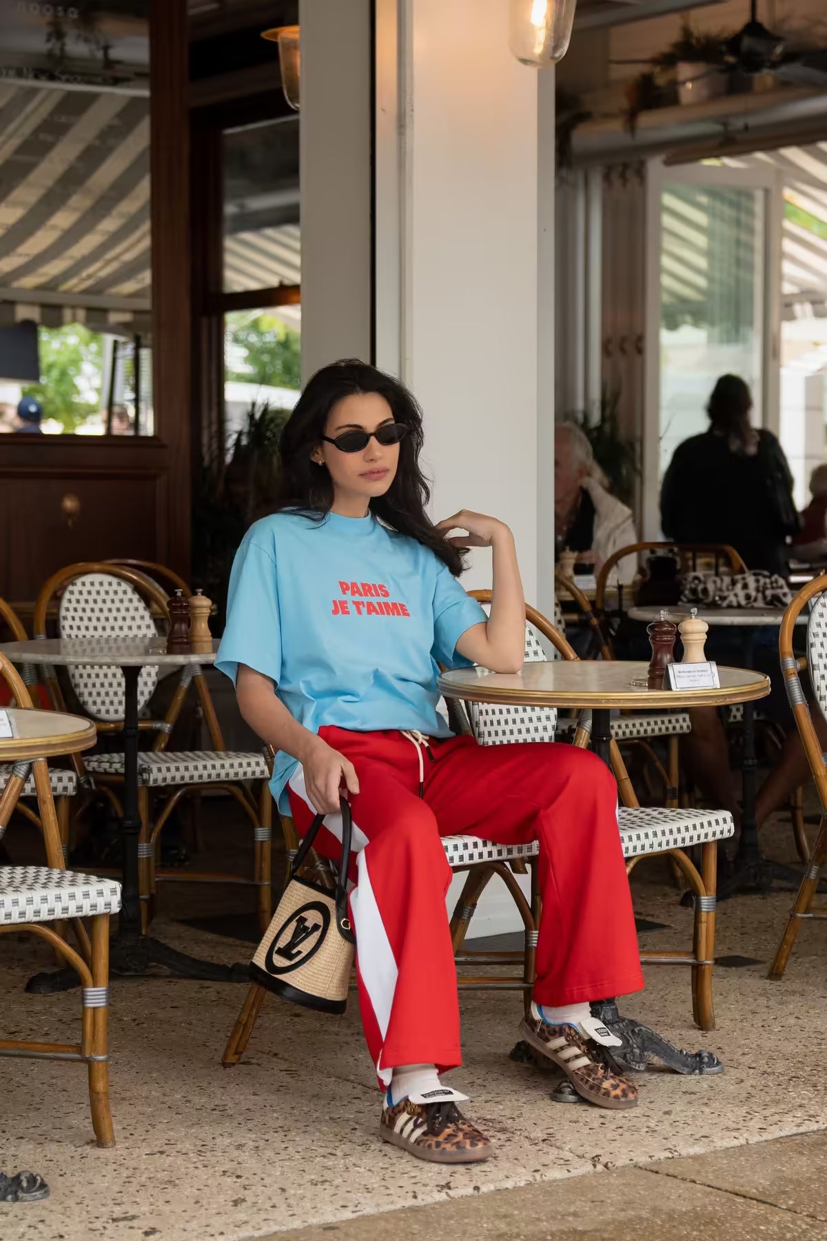 Woman sitting at an outdoor cafe wearing a blue shirt with red text and red pants.