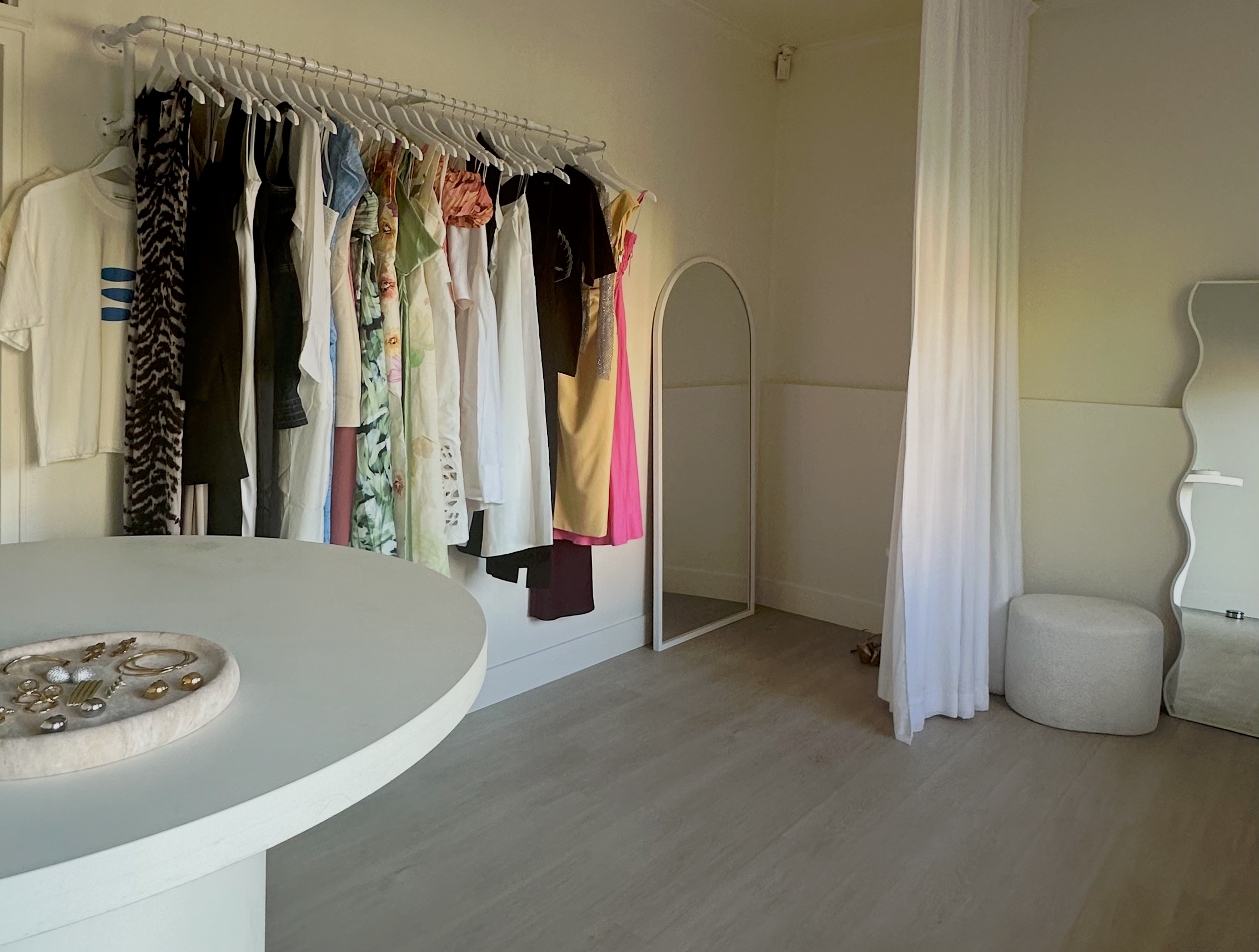 A boutique fitting room with a clothing rack filled with garments, a white round table in the foreground, a full-length mirror, a white curtain, and a round ottoman on light wood flooring.