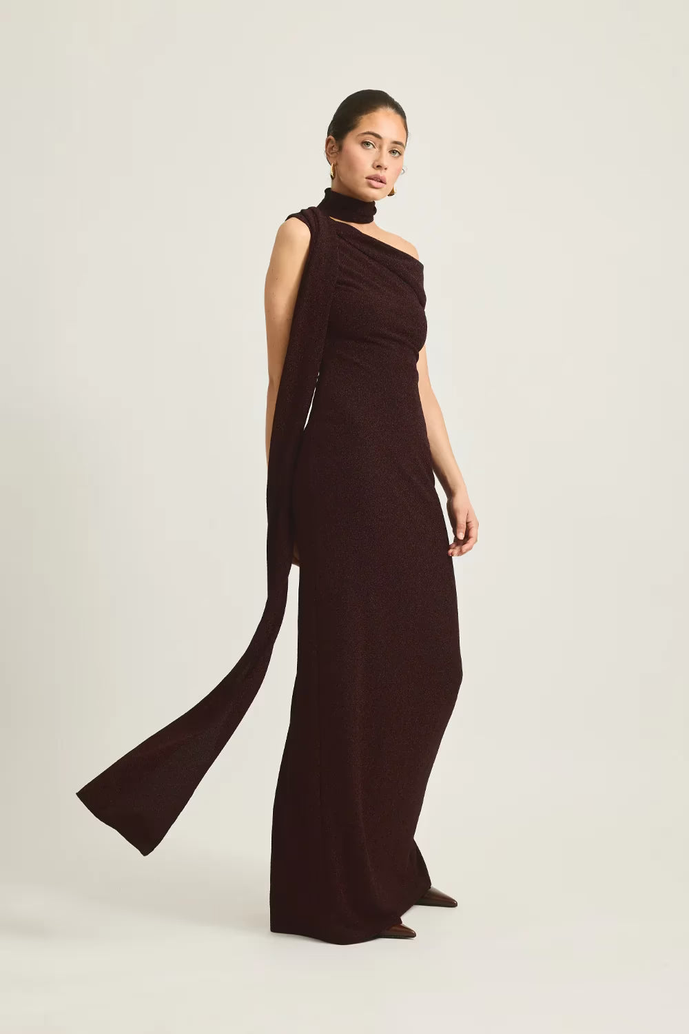 Izara dress in deep burgundy, featuring a one-shoulder design and a flowing fabric drape, worn by a model.