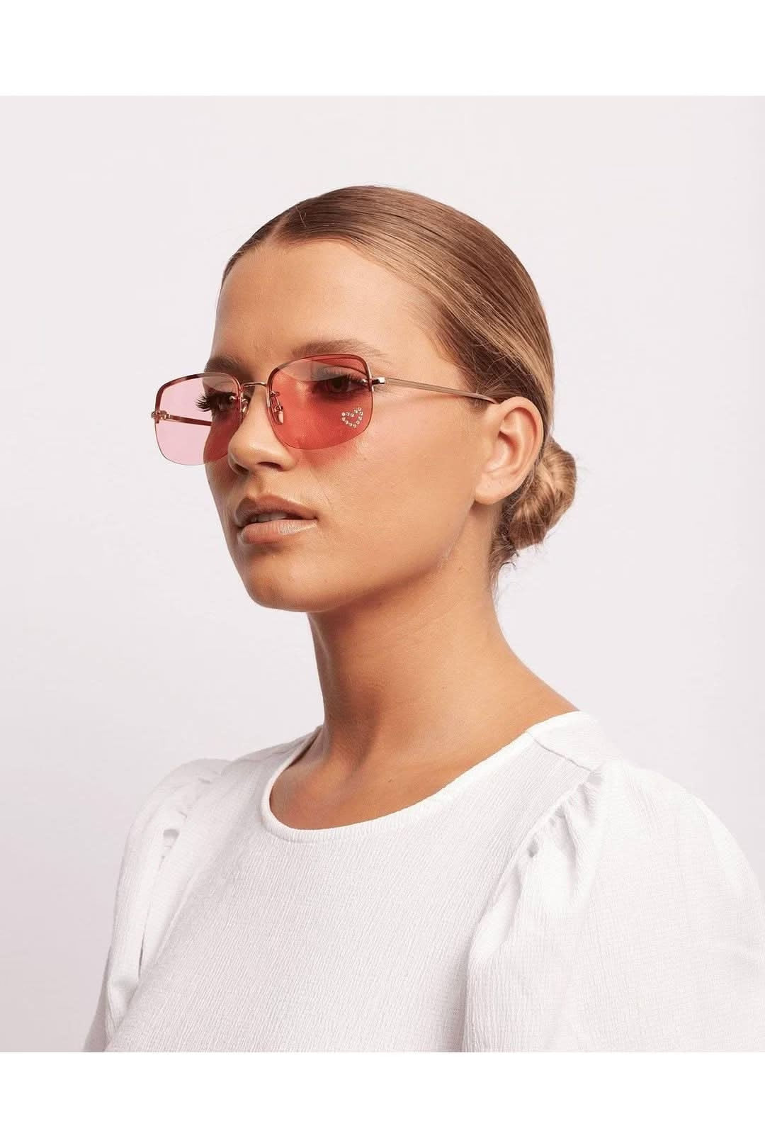 Faster Love sunglasses with pink lenses, worn by a model in a white blouse against a light background.