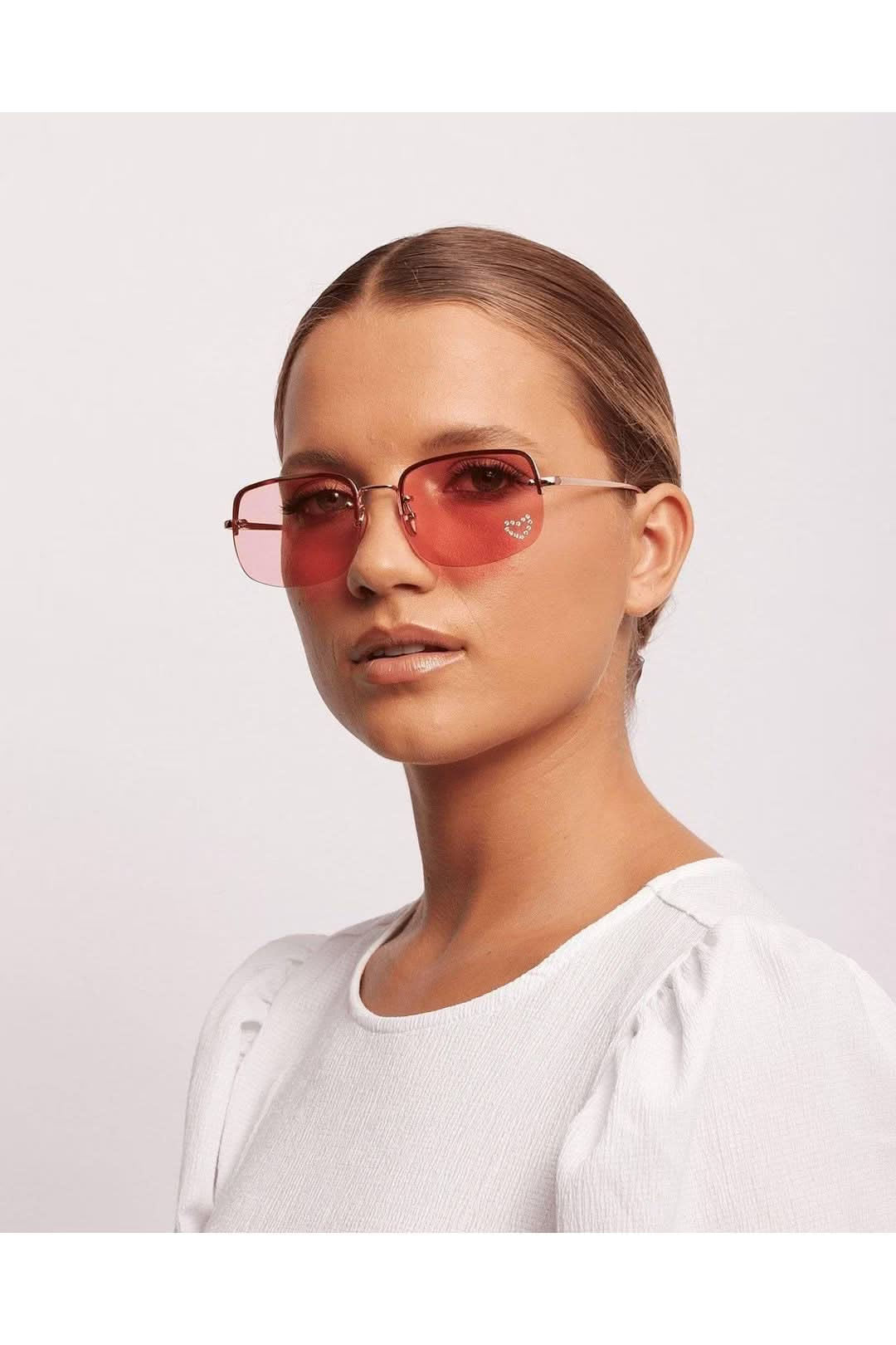 Faster Love sunglasses with pink lenses and gold frames, worn by a model in a white top against a neutral background.
